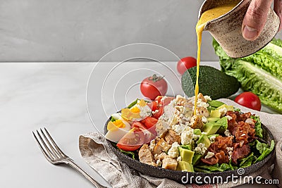 A hand pouring salad dressing into cobb salad with chicken, avocado, bacon, blue cheese, tomato and eggs. Keto diet Stock Photo