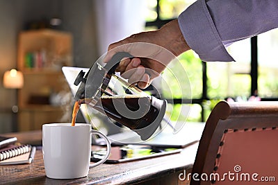 A hand pouring steaming coffee in to a cup on a work desk when work from home Stock Photo