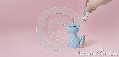 Hand puts a dollar in the kitty`s piggy Bank corn blue with a colorful rainbow tail and unicorn horn on a pink Stock Photo