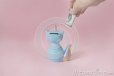 Hand puts a dollar in the moneybox kitten corn blue with a colorful rainbow tail with closed eyes and a unicorn horn on Stock Photo