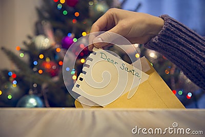 A hand putting a paper with Dear Santa inscription into an envelope on a wooden table against decorated Christmas tree Stock Photo