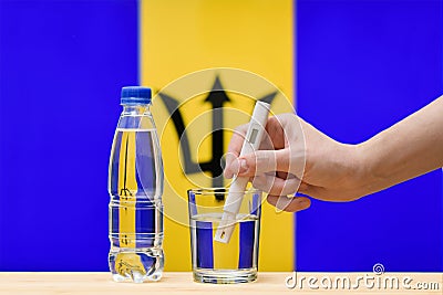 A hand with a water tester makes a measurement in a glass of clear water against the background of the flag of Barbados. Stock Photo