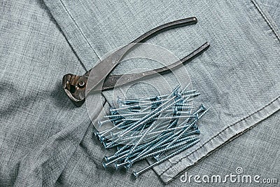 A handful of metallic self-cutters screws and very aged and rusty pliers lies on backdrop of an old denim fabric Stock Photo