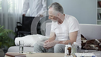 Handicapped man sitting on sofa at nursing home, male nurse bringing wheelchair Stock Photo