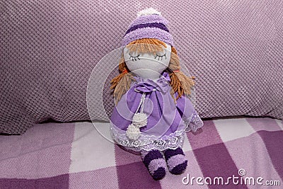 Handmade doll in purple hat and dress on sofa in room. Stock Photo