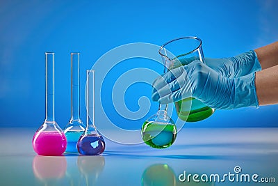 Hands in disposable gloves pouring green liquid from beaker into medical flask. Colorful chemical reagents, blue Stock Photo