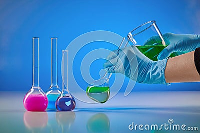 Hands in disposable gloves pouring green liquid from beaker into medical flask. Purple, pink chemical reagents, blue Stock Photo