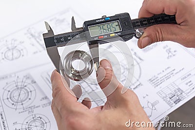 Hands of an engineer measures a metal part with a digital vernier caliper against the background of technical drawings. Quality Stock Photo