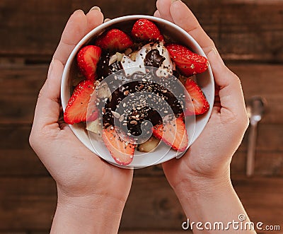 Hands holding strawberries with yogurt chocolate pumpkin seeds chia sunflower seeds and apple in a white bowl on a wooden table Stock Photo