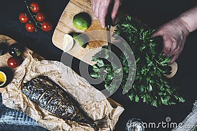 Hands in the process of cooking fish, pepper, parsley, tomato, lime on a cutting board on a black wooden background top view horiz Stock Photo