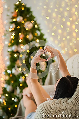 Hands in the shape of a heart on the background of the Christmas tree. Girl on sofa cozy heart background. Hygge style with yello Stock Photo