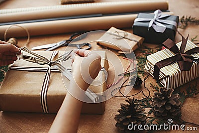 Hands wrapping stylish christmas gift box in craft paper and scissors, rustic presents, thread, pine branches and cones on wooden Stock Photo