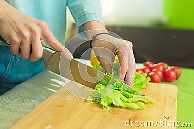 Hands of a young girl slice green lettuce leaves on a wooden cutting board on a green table in a home setting against a Stock Photo
