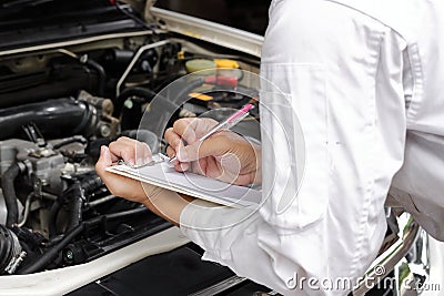 Hands of young professional mechanic in uniform writing on clipboard against car in open hood at the repair garage. Maintenance se Stock Photo