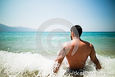 Handsome attractive muscular man sitting on sea shore on the beach sand and relaxing.Handsome man with tattoo sunbathing, spf Stock Photo