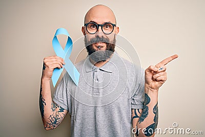 Handsome bald man with beard and tattoos holding blue cancer ribbon over isolated background very happy pointing with hand and Stock Photo