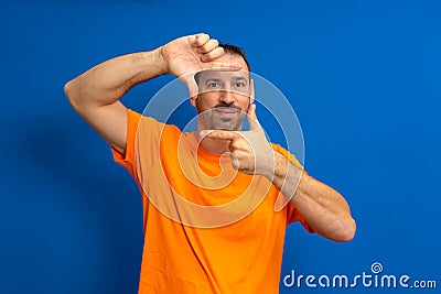 Handsome bearded happy male blinks his eyes and looks through a frame formed by his hands. An attractive man makes a Stock Photo