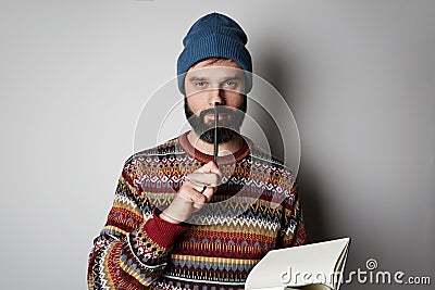 Handsome bearded man in blue beanie thinking with pen and notebook over background. Stock Photo