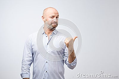 Handsome bearded man in casual clothes is pointing awaywith thumb looking at camera on gray background Stock Photo