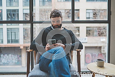 Handsome bearded man using mobile phone and drinking coffee at home. Casual relaxing and rest time concept. Blurred Stock Photo