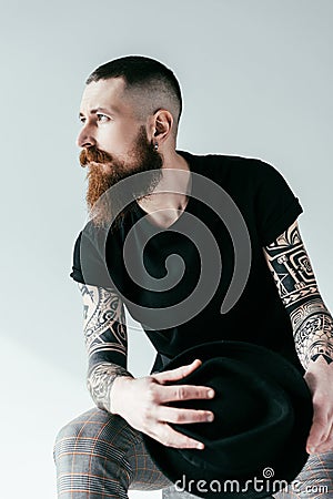 handsome bearded tattooed man holding hat and looking away Stock Photo