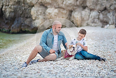 Handsome bold stylish dressed in jeans man in blue cap sitting on a beach sea side together with cute son boy teen andpretty engli Stock Photo