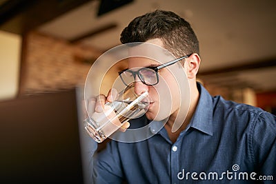 Handsome caucasian man with glass of water in hand works with laptop. Businessman in glasses drinks water for body Stock Photo