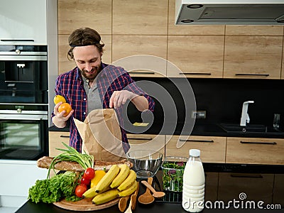 Handsome Caucasian man unpacking purchased healthy food standing by a kitchen countertop with bottle of milk and healthy raw vegan Stock Photo