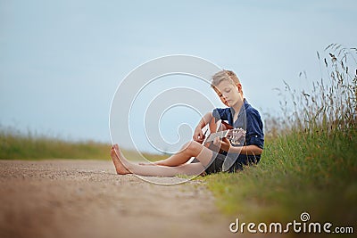 Handsome cute boy is playing on acoustic guitar sittingon road in summer day. Stock Photo