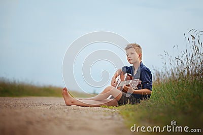 Handsome cute boy is playing on acoustic guitar sittingon road in summer day. Stock Photo