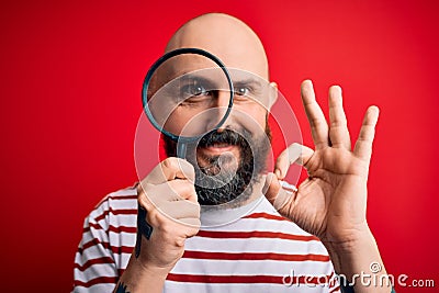 Handsome detective bald man with beard using magnifying glass over red background doing ok sign with fingers, excellent symbol Stock Photo