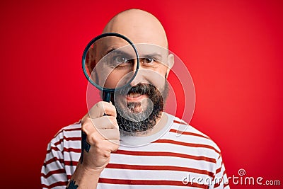 Handsome detective bald man with beard using magnifying glass over red background with a happy face standing and smiling with a Stock Photo