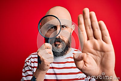 Handsome detective bald man with beard using magnifying glass over red background with open hand doing stop sign with serious and Stock Photo