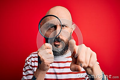 Handsome detective bald man with beard using magnifying glass over red background pointing with finger to the camera and to you, Stock Photo