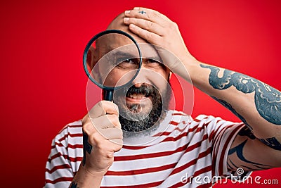 Handsome detective bald man with beard using magnifying glass over red background stressed with hand on head, shocked with shame Stock Photo