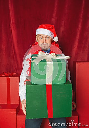 Handsome funny man wearing Santa hat holding heavy Christmas gift boxes on the red background. New Year style Stock Photo