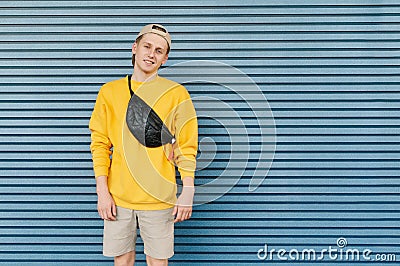 Handsome guy in stylish streetwear stands on the background of a blue wall,smiles and looks at the camera, wears a cap and a fanny Stock Photo