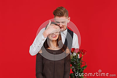 The guy from behind closes the girl`s hand to her eyes with a bouquet of roses on a red background Stock Photo