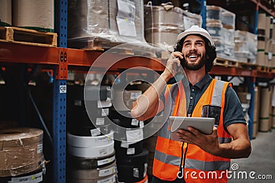 Handsome happy man in white helmet and safety vest standing holding digital tablet while talking over phone looking away Stock Photo