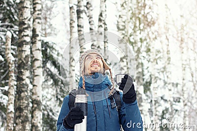 Handsome hiker man in warm clothes with thermos travel in winter forest in snowy day. He looks up and smile. Season concept. Copy Stock Photo