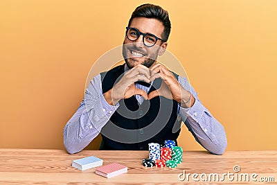Handsome hispanic croupier man sitting on the table with poker chips and cards smiling in love showing heart symbol and shape with Stock Photo