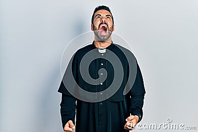Handsome hispanic man with beard wearing catholic priest robe angry and mad screaming frustrated and furious, shouting with anger Stock Photo