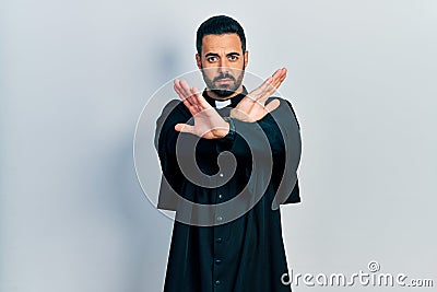 Handsome hispanic man with beard wearing catholic priest robe rejection expression crossing arms and palms doing negative sign, Stock Photo