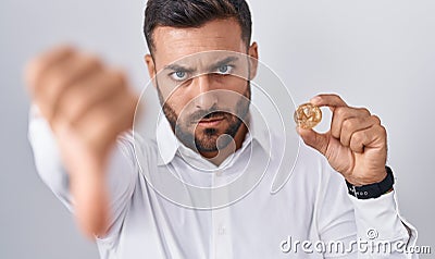 Handsome hispanic man holding monero cryptocurrency coin with angry face, negative sign showing dislike with thumbs down, Editorial Stock Photo