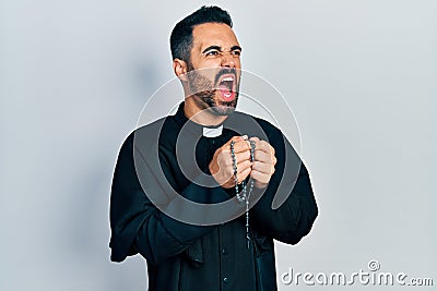 Handsome hispanic priest man with beard praying holding catholic rosary angry and mad screaming frustrated and furious, shouting Stock Photo