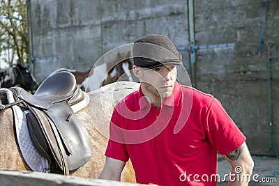 Handsome horse rider in red polo shirt and flatcap standing next to his saddled horse Stock Photo
