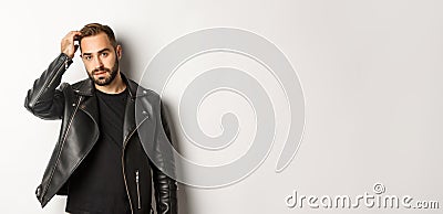 Handsome macho man in black biker jacket, touching his haircut and looking cool, white background Stock Photo