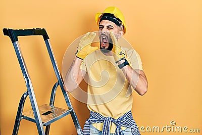 Handsome man with beard by construction stairs wearing hardhat shouting angry out loud with hands over mouth Stock Photo