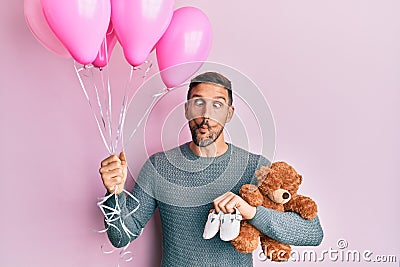 Handsome man with beard expecting a baby girl holding balloons, shoes and teddy bear making fish face with mouth and squinting Stock Photo