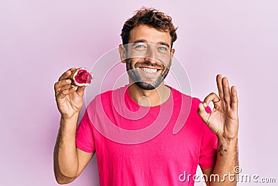 Handsome man with beard holding pink geode precious gemstone doing ok sign with fingers, smiling friendly gesturing excellent Stock Photo